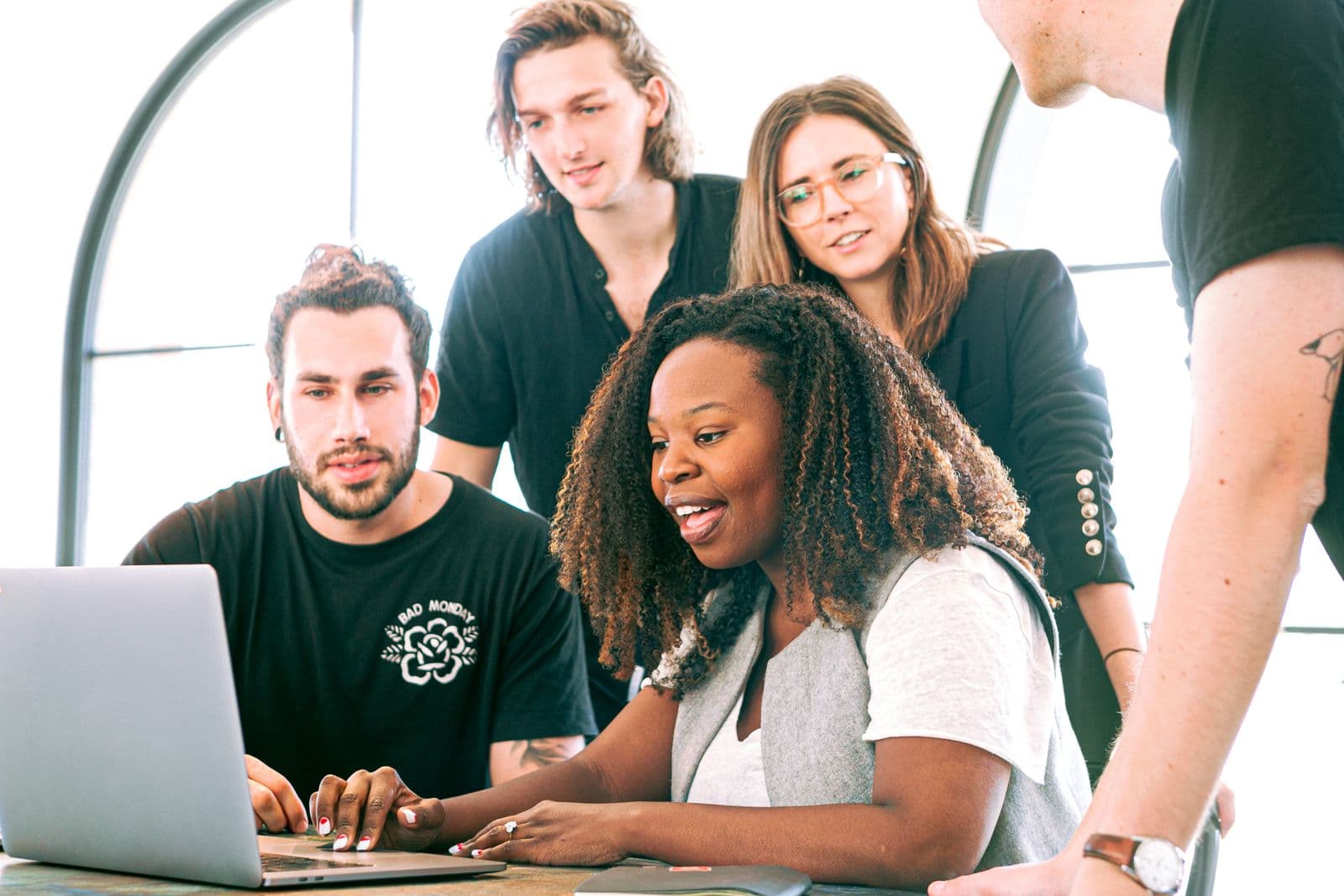 Four people working together at a laptop beside a window.