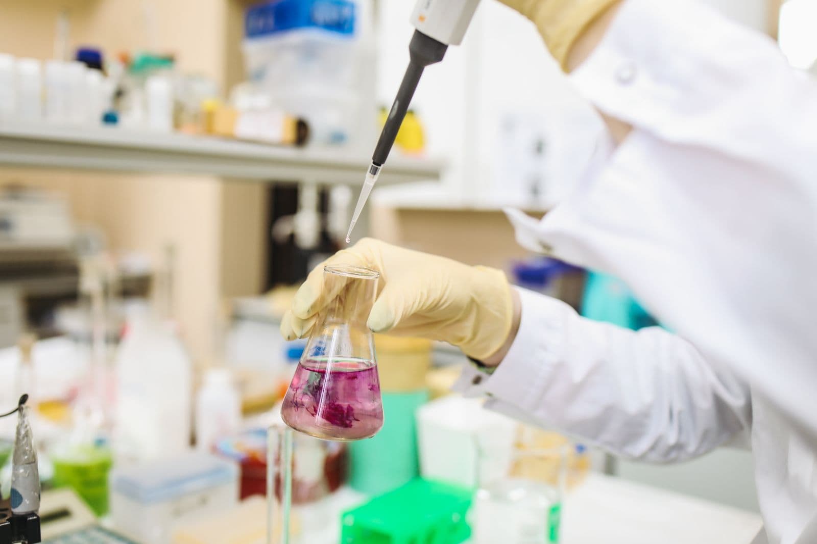 A gloved hand using a pipette over sample flasks in a laboratory.