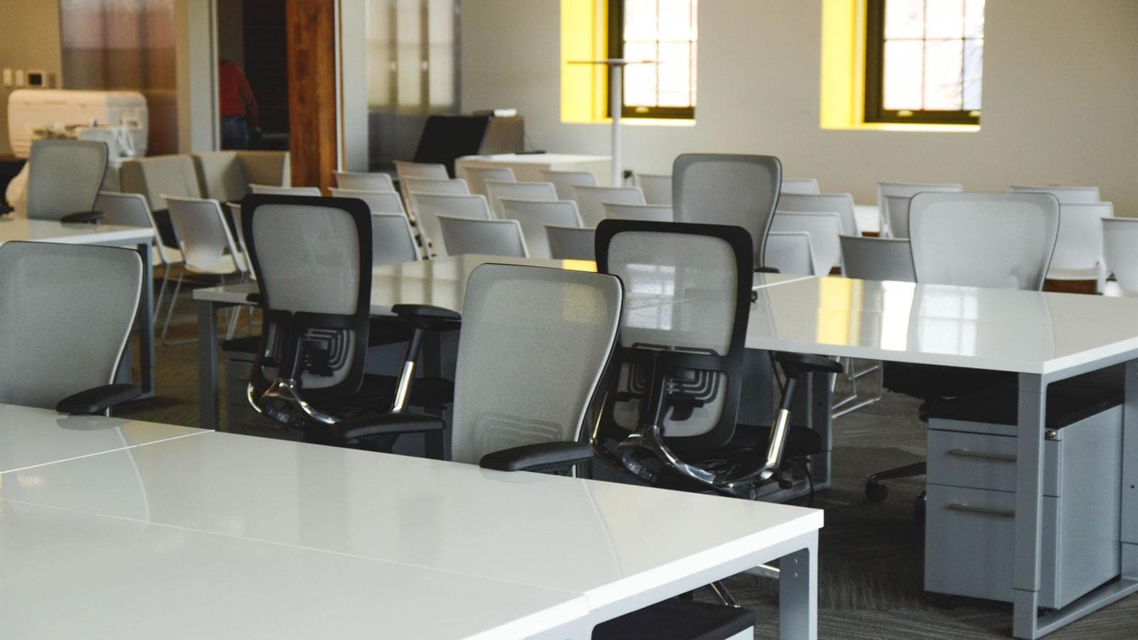 An open workspace with flexible desks and task chairs, lit by tall windows.
